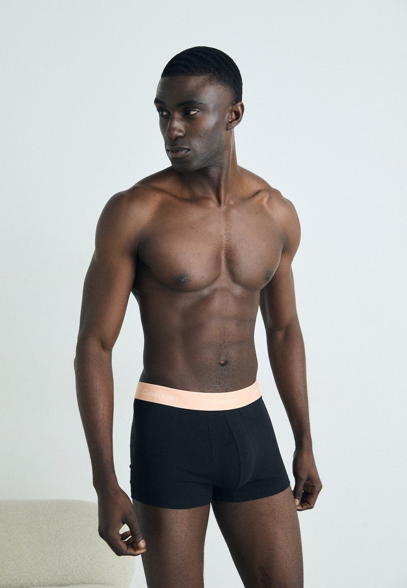 Boxer-briefs noirs avec une ceinture Calvin Klein pêche clair, portés par un mannequin masculin aux cheveux courts et ondulés, sur un fond neutre.
