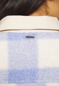 Fuzzy fleece jack met een lichtblauw en wit geblokt patroon. Bevat een beige kraag en een metallic Billabong-logo-tag.