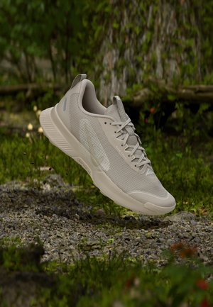 Pantof de alergare trail Nike bej deschis suspendat deasupra terenului stâncos, cu vegetație verde și trunchi de copac în fundal.