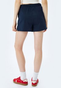 Marineblaue Shorts mit einem elastischen Bund, Seitentaschen und einer Gesäßtasche, aus glattem Stoff. Getragen mit roten Sneakers und weißen Socken.