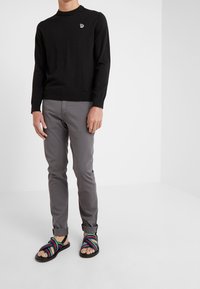 Pull-over noir, pantalon gris ajusté et sandales multicolores à brides. Détails notables : un logo brodé sur le pull.