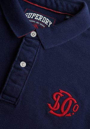 Marineblauw Superdry poloshirt met rood geborduurd logo, witte knopen en een rood stikseldetail op de kraagrand.