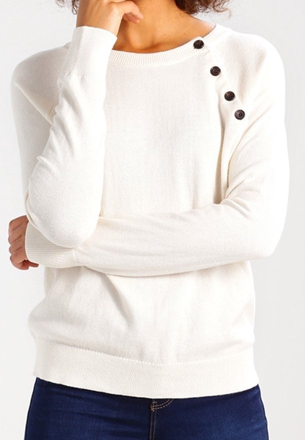 Femme portant un pull blanc à manches longues avec trois boutons noirs sur l'épaule gauche, associé à un jean bleu foncé.
