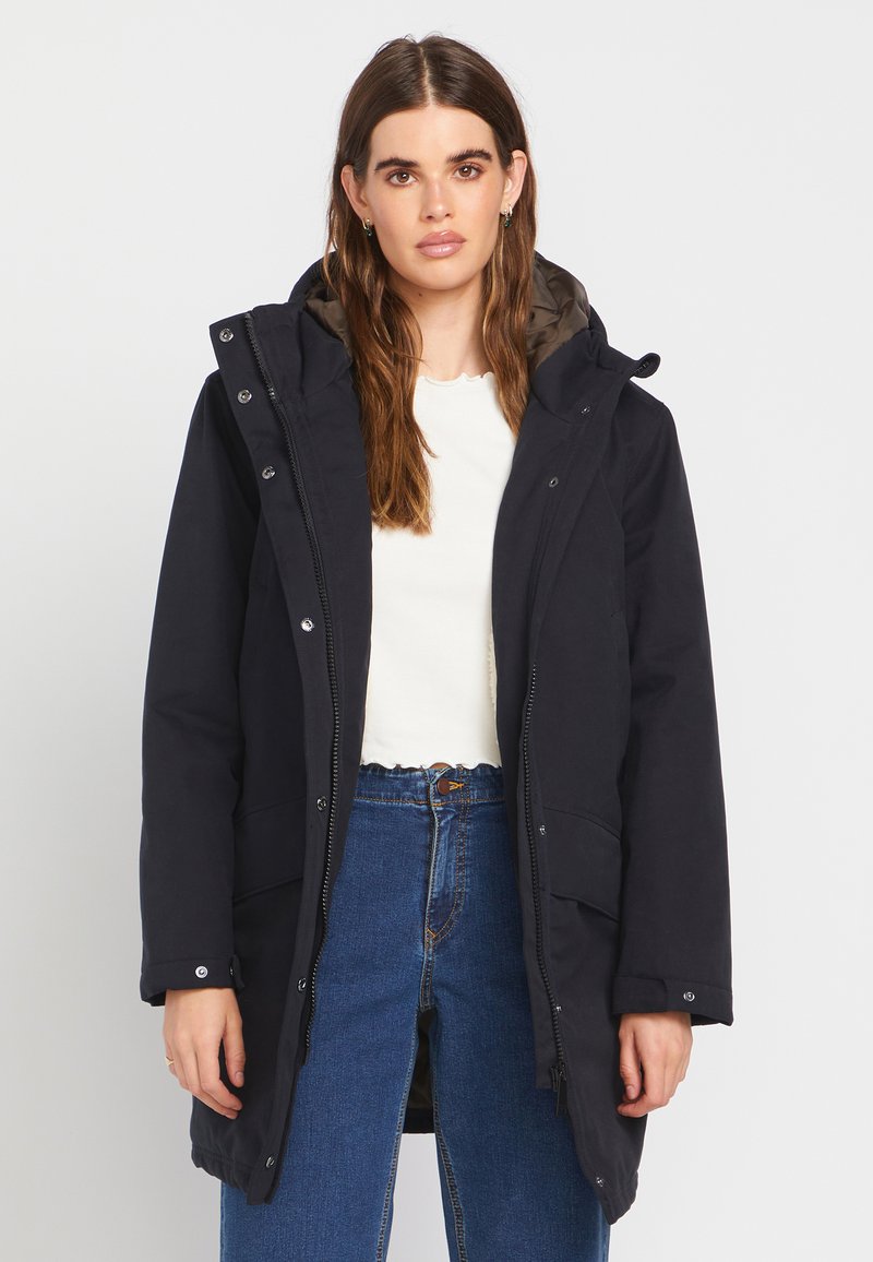 Femme aux longs cheveux bruns portant un manteau d'hiver noir à capuche, un haut blanc et un jean bleu, debout devant un fond clair uni.