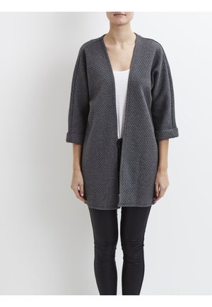 Gilet gris ouvert avec un motif de tricot texturé, des manches trois-quarts et une coupe décontractée, porté sur un top blanc et un legging noir.