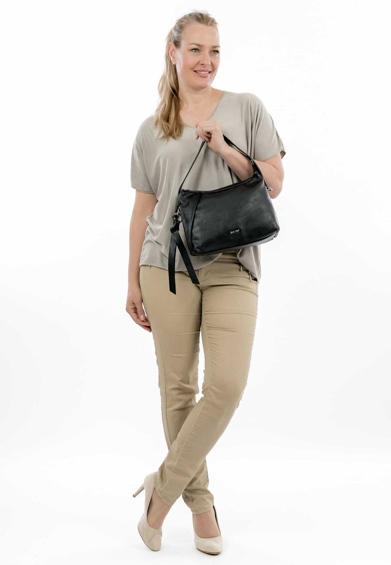 Musta nahkakäsilaukku, jossa on tasainen pinta, yksi hihna ja vetoketjukiinnitys. Malli, joka on pukeutunut neutraaleihin väreihin, seisoo rentona.