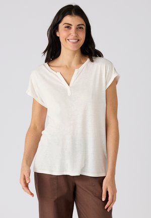 Lachende vrouw met donker haar, gekleed in een wit kortemouwen blouse met een ingesneden hals en een bruine broek, staand tegen een effen achtergrond.