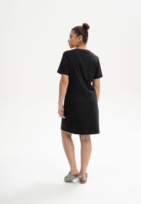 Schwarzes Baumwoll-T-Shirt-Kleid mit kurzen Ärmeln, lockerem Sitz, bis zur Mitte des Oberschenkels reichend, kombiniert mit grauen offenen Clogs.