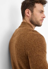 Pull marron tricoté en fil texturé, avec un col montant et des manches raglan, présenté de profil sur un fond clair.