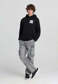 Felpa nera con stampa grafica, abbinata a pantaloni cargo grigi chiari con numerose tasche. Sneakers nere con suola bianca completano il look.