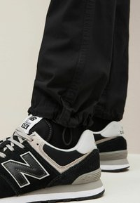 Svarta New Balance-sneakers med grå detaljer, med ett överdel i mesh och mocka samt en vadderad sula; kombinerade med svart byxor med elastiskt botten.