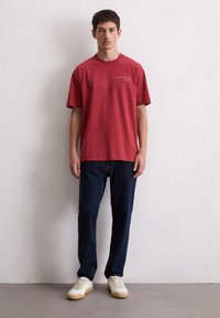 Rotes Baumwoll-T-Shirt mit kurzen Ärmeln, Rundhalsausschnitt und weißem Text auf der Vorderseite; kombiniert mit dunkelblauen Jeans und hellen Turnschuhen.