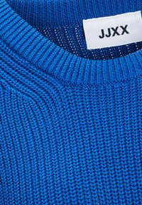 Pull bleu en tricot avec un col rond côtelé et une étiquette en tissu blanc portant l'inscription « JJXX » cousue à l'intérieur du col.