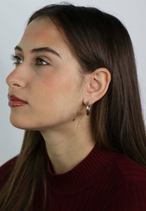 Young woman with long brown hair wearing silver hoop earrings and a burgundy ribbed top, looking calmly to the left against a plain background.