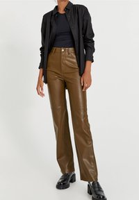 Un pantalon évasé en cuir marron présente une taille haute et des coutures frontales, associé à une chemise noire à boutons. Des chaussures à plateforme noires viennent compléter le look.
