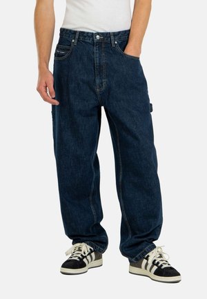 Pantalones vaqueros baggy de mezclilla de color azul oscuro con una cintura alta, bolsillos frontales y costuras visibles. Combinados con zapatillas negras y blancas.