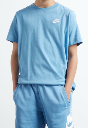 Osoba ubrana w pasującą jasnoniebieską koszulkę i spodenki Nike z białymi logo Nike, ręce w kieszeniach, widoczna od szyi do połowy uda.