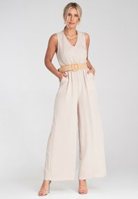 Beżowy kombinezon bez rękawów z dekoltem w serek, szerokimi nogawkami i teksturowanym paskiem. Posiada boczne kieszenie oraz gładki, lejący się materiał.
