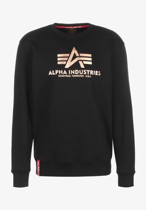 Sweat-shirt noir en coton, avec un logo métallique doré "ALPHA INDUSTRIES" orné d'un design triangulaire, et une étiquette rouge sur l'ourlet.