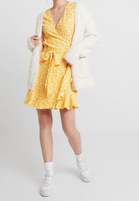Robe portefeuille à fleurs jaunes avec un ourlet volanté et une taille à nouer, associée à un cardigan blanc duveteux. Des baskets blanches et des chaussettes montantes complètent le look.