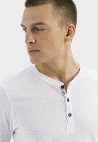 Weißes, langärmeliges Henley-Shirt aus weichem Material mit zwei schwarzen Knöpfen und einem kleinen Druckdetail am Ausschnitt. Zarte Textur sichtbar.