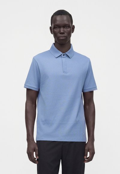 Homme debout portant un polo bleu clair texturé à manches courtes avec un pantalon noir sur un fond clair uni.