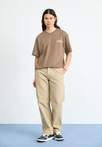 Brun bomull T-skjorte med korte ermer og logo på brystet, sammen med khaki cargo-bukser. Svart-hvite joggesko med teksturert overdel.