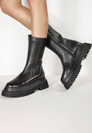 Jambes portant des bottes en cuir noires mi-mollet avec des semelles épaisses et robustes et des fermetures éclair latérales, positionnées sur un fond blanc uni.