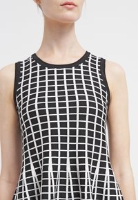 Femme portant une robe noire sans manches avec un motif à grille blanche, debout devant un fond clair uni.