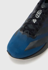 Kék futócipő texturált felsőrésszel, szürke részletekkel, elegáns kialakítással és fekete fűzőkkel. Jellegzetes logó látható az oldalán.