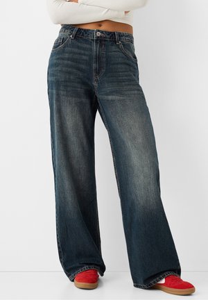 Person iført mørke vide jeans, en cropped hvid langærmet top og røde sneakers står mod en ensfarvet hvid baggrund.