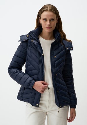 Giacca puffer blu navy con chiusura a zip, dettagli in metallo dorato, motivo a zig-zag, cappuccio removibile e polsini elasticizzati, indossata sopra un top crema.