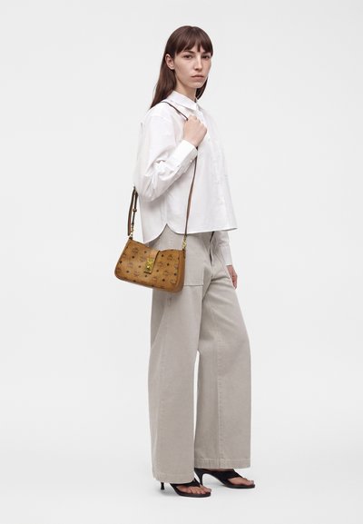 Femme portant une chemise blanche et un pantalon large beige tient un sac à main marron avec une fermeture dorée, debout devant un fond uni.