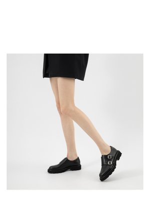 Osoba nosząca czarne mokasyny z podwójną klamrą i czarną spódnicę, lekko unosząca jedną stopę nad ziemię na jednolitym tle.