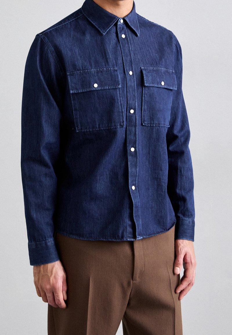 Homme portant une chemise en denim bleu foncé boutonnée avec deux poches poitrine et un pantalon marron, debout devant un fond uni.