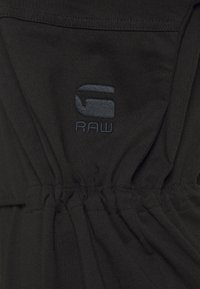 Svart tygtröja med volangdetaljer vid midjan, med en mörkgrå broderad logotyp "G RAW" på framsidan.