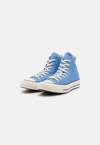 Sneaker alte, tomaia in tela azzurra chiara, lacci bianchi, suola in gomma con striscia nera, punta tonda e dettagli di cucitura.