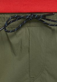 Olivgröna shorts med en texturerad midja i rött, som har en svart dragsko med blå detaljer och synliga sömnadsdetaljer.