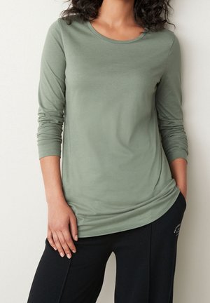 Femme portant une chemise vert clair à manches longues et un pantalon noir, debout avec une main dans la poche et l'autre reposant sur la cuisse.