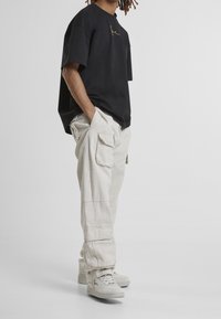 Henkilö, jolla on päällään musta oversized t-paita, vaaleanbeige cargo-housut sivutaskuilla ja valkoiset lenkkarit, seisoo yksinkertaista taustaa vasten.