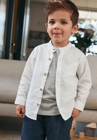 Camicia di lino bianca con bottoni di legno, indossata sopra una t-shirt grigia, abbinata a pantaloni blu scuro. Tessuto liscio e design casual.