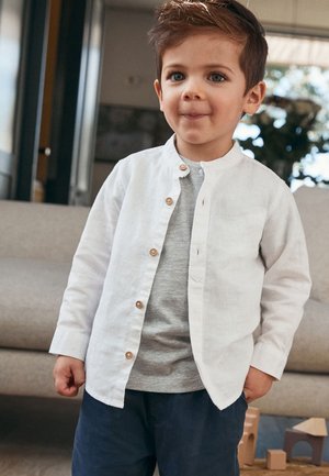 Weiße Leinenbluse mit Holzknöpfen über einem grauen T-Shirt, kombiniert mit dunkelblauen Hosen. Glatte Textur und lässiges Design.