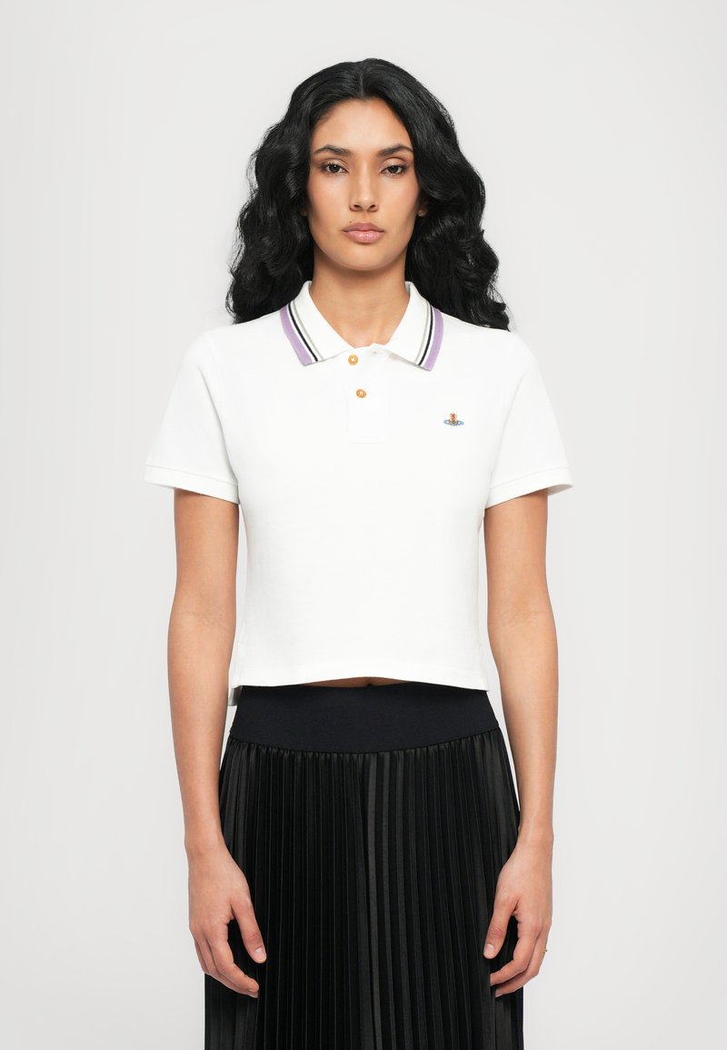 Hvid cropped polo skjorte med lilla og sorte striber i kraven, to orange knapper og broderet logo. Kombinationen med en plisseret sort nederdel.