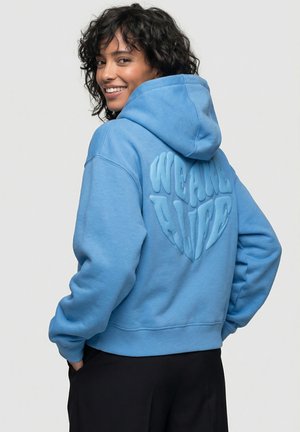 Frau lächelt, trägt einen blauen Hoodie mit erhabenen Buchstaben "WE ARE ALIVE", die auf dem Rücken die Form eines Herzens bilden, und steht mit den Händen in den Taschen.