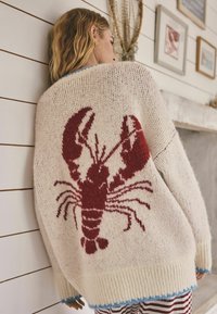 Džemper pleteni u boji slonovače s velikim crvenim motivom jastoga. Ima rebraste manžete i plavi obrub uz rub. Mekani, teksturirani materijal.