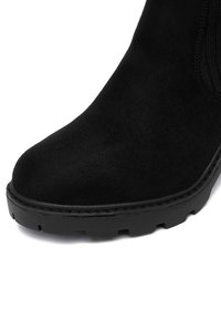 Fekete velúr bokacsizma lekerekített orral, texturált felülettel és vastag gumitalppal, amelynek barázdái biztosítják a tapadást. Varrott részletek láthatóak.