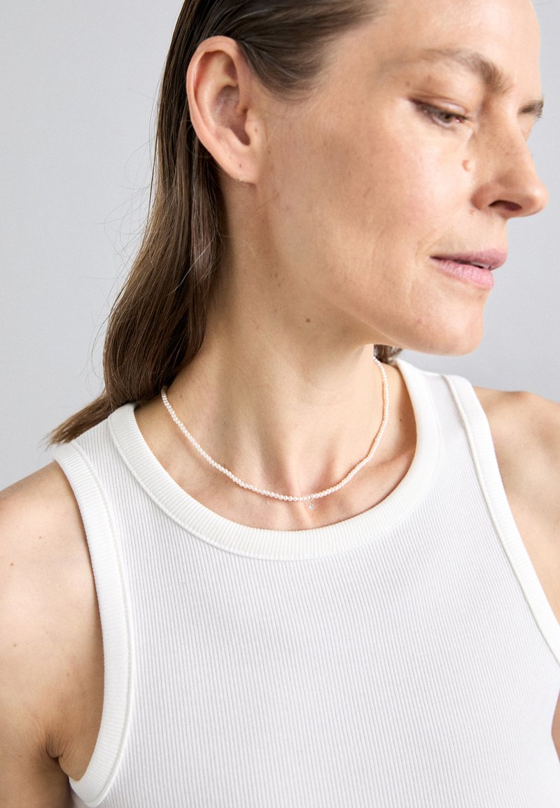 Pērļu kaklarota ar delikātām, apaļām pērlēm un nelielu piekariņu, uzvilkta virs teksturēta baltā topa un akcentēta ar maigu dabisku apgaismojumu.