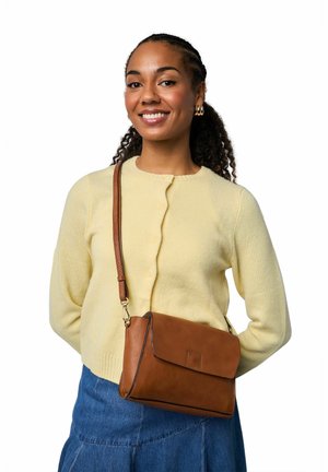 Sac bandoulière en cuir marron avec fermeture à rabat ; texture douce et forme structurée. Porté avec un cardigan jaune clair et une jupe en jean.