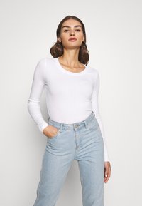 Weiße, gerippte Langarmbluse mit rundem Ausschnitt, kombiniert mit hellblauen, hoch geschnittenen Jeans. Glatte Textur, taillierter Schnitt, ohne Muster.