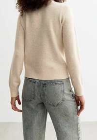 Beige gestrickter Pullover mit geripptem Saum und langen Ärmeln, kombiniert mit ausgewaschenen blauen hochgeschnittenen Jeans mit doppelten Gesäßtaschen.